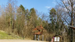 Eine Drachenstatue steht in einer Waldlichtung unter blauem Himmel. Der Boden ist mit Kies bedeckt. Bäume umgeben das Gebiet, und ein Weg führt zu einem Grashügel.