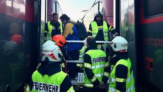 Feuerwehrleute in neongrünen Uniformen und Helmen helfen einem Mann mit Rucksack, möglicherweise auf einem Bahnsteig.