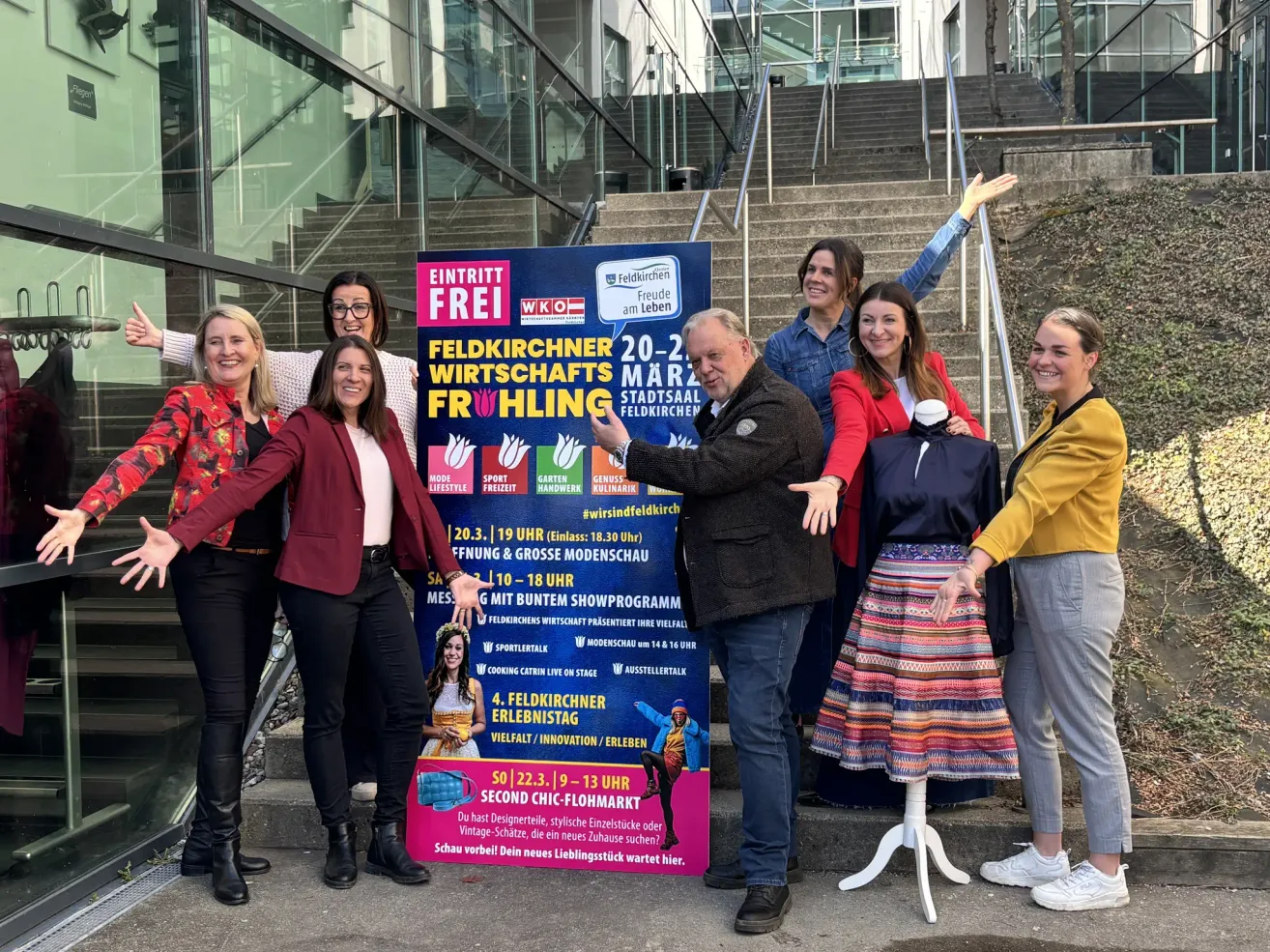 Eine Gruppe von Menschen steht vor einem Schild für eine Modeveranstaltung. Die Veranstaltung heißt Feldkirchner Wirtschaftsfruhling. Das Schild hat einen Zeitplan und eine Liste von Aktivitäten.