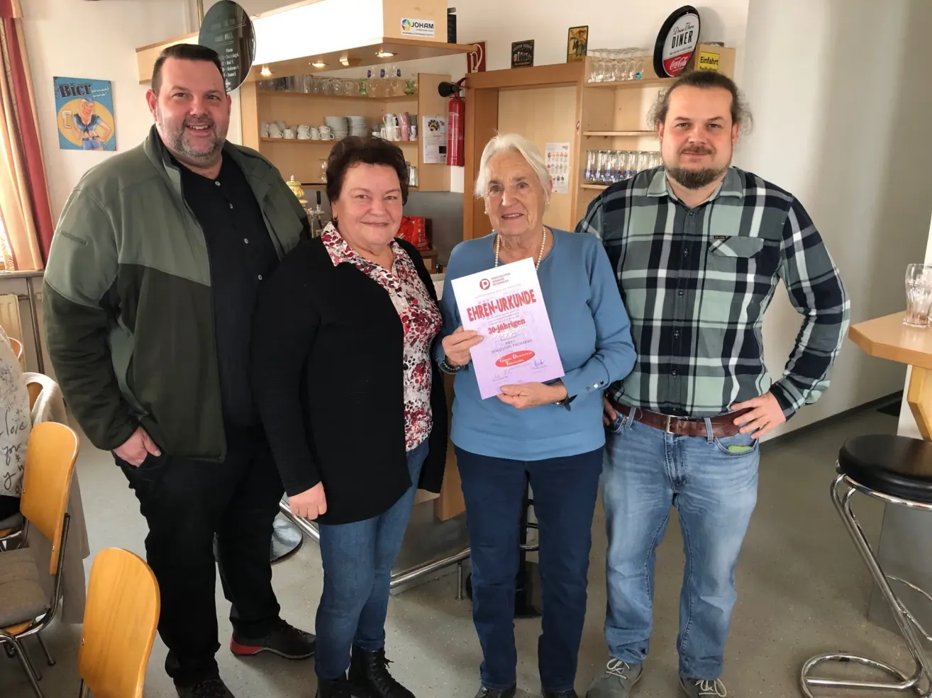 Vier Personen, darunter eine Frau, die ein Zertifikat in der Hand hält, posieren für ein Foto. Sie stehen eng beieinander in einem Raum. Hinter ihnen steht ein Regal mit verschiedenen Gegenständen.