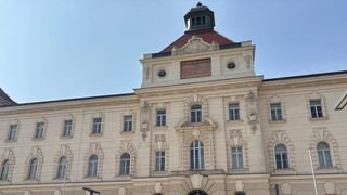 Ein großes weißes Gebäude mit vielen Fenstern und einem Turm mit einem braunen Dach. Ein Schild steht 'Kaiserlicher Hof'.