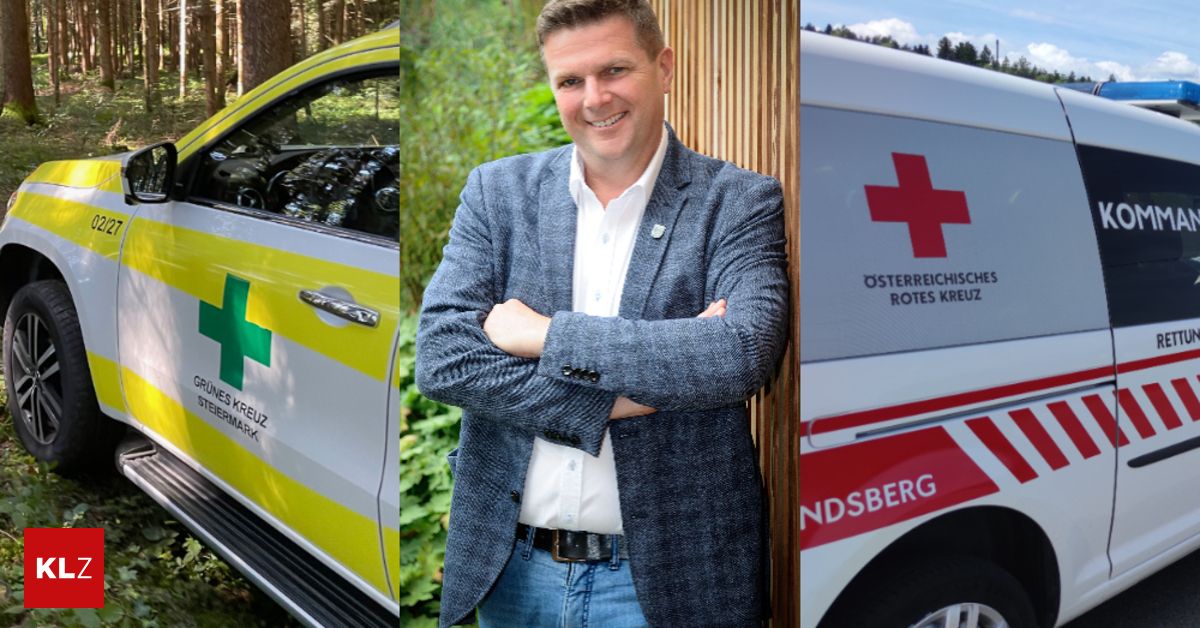 Ein lächelnder Mann mit verschränkten Armen steht zwischen einem Krankenwagen und einem Auto, alles vor einem grünen Hintergrund.