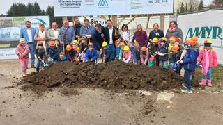 Eine Gruppe von Menschen, darunter Erwachsene und Kinder, halten Schaufeln für eine feierliche Grundsteinlegung. Sie tragen orangefarbene Schutzhelme und gelbe Jacken. Hinter ihnen steht ein Schild mit der Aufschrift NO Landeskindergarten und Tagesbetreuung Ulmerfeld-Hausmening.