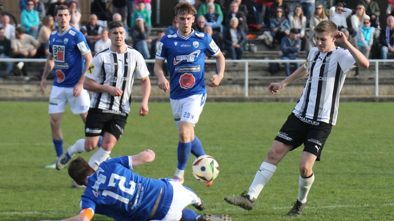 Fußballspieler in blauen und weißen Trikots spielen auf einem Feld. Ein Spieler in Blau läuft, während ein anderer in Weiß fällt. Ein Publikum sieht von den Rängen zu.