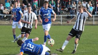 Fußballspieler in blauen und weißen Trikots spielen auf einem Feld. Ein Spieler in Blau läuft, während ein anderer in Weiß fällt. Ein Publikum sieht von den Rängen zu.