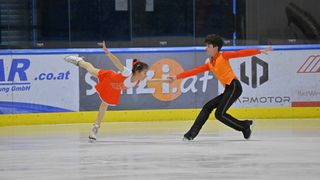 Zwei junge Eiskunstläufer, ein Junge und ein Mädchen, führen eine Routine auf der Eisbahn durch, wobei das Mädchen in der Luft ist. Dahinter befindet sich eine Wand mit Werbeanzeigen.