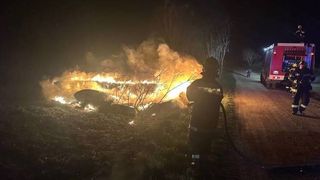 Feuerwehrleute löschen ein brennendes Auto auf einer Landstraße in der Nacht. Das Auto ist vollständig in Flammen aufgegangen, mit Feuerwehrleuten in der Nähe.
