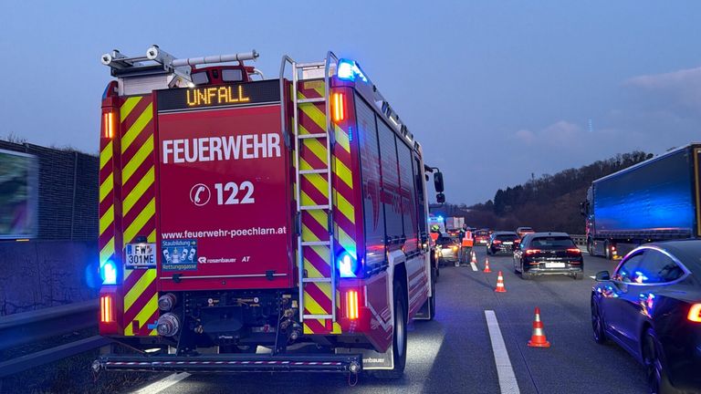 Ein roter Feuerwehrwagen mit 'Unfall' und 'Feuerwehr 122', Lichter an und eine Leiter. Autos, Kegel und Menschen mit Westen auf der Straße.