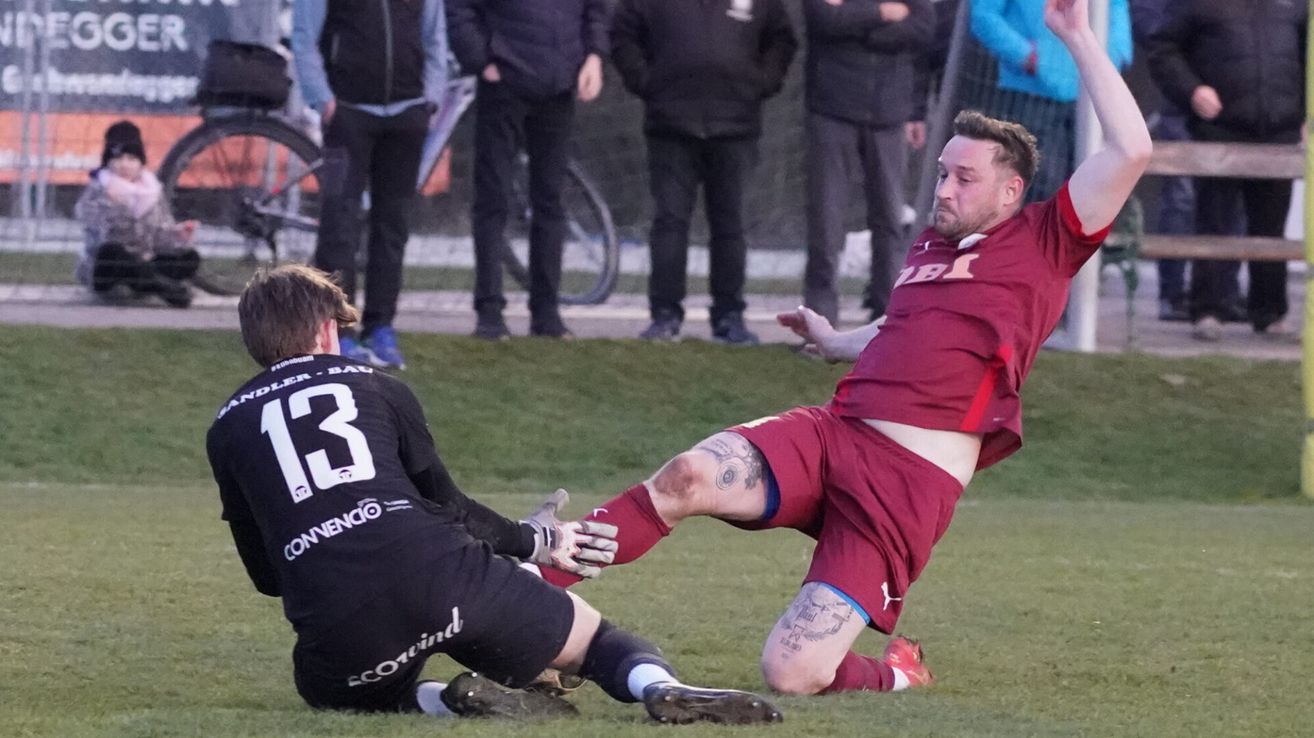 Ein Fußballspieler in einem Weinrot-Trikot fällt zu Boden, während ein anderer Spieler versucht, ihn zu blockieren. Zuschauer stehen hinter einem Zaun und beobachten.
