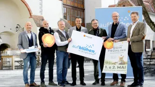 Sechs Männer stehen vor einem Gebäude mit Bannern. Einer hält ein Banner mit der Aufschrift 'aufbluehen'. Ein anderer hält ein Sonnenzeichen. Ein Schild sagt 'Wein Frühling' mit den Daten 1. und 2. Mai.