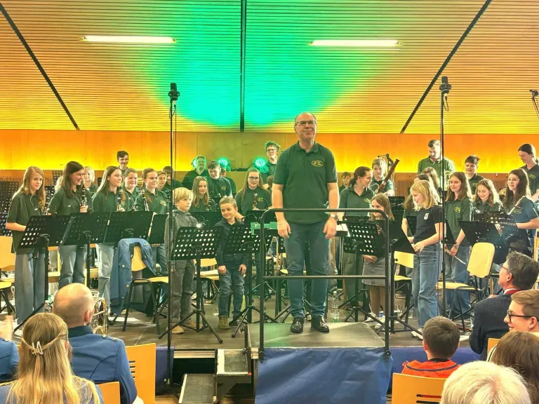 Ein Mann steht auf einer Bühne mit einer Gruppe von Kindern in grünen Hemden. Sie spielen Instrumente und das Publikum schaut zu.