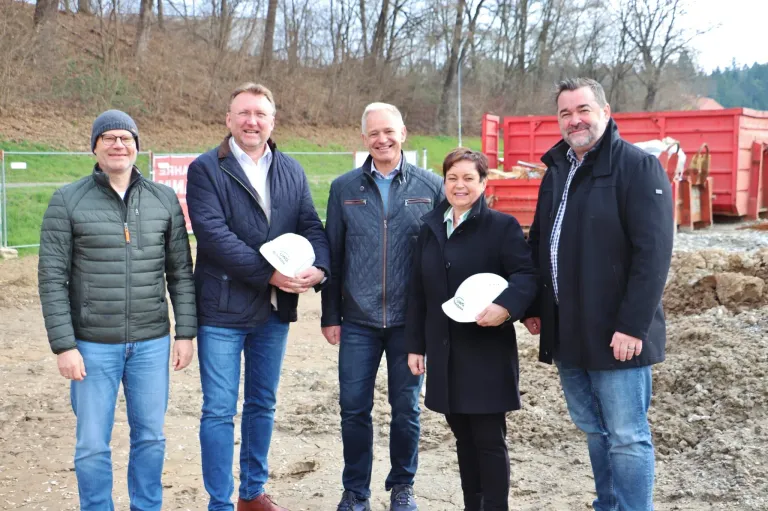 Fünf Personen stehen lächelnd in einer Gruppe und halten weiße Mützen. Hinter ihnen befindet sich ein Rasen mit Bäumen und einem roten Container.