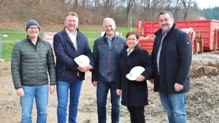 Fünf Personen stehen lächelnd in einer Gruppe und halten weiße Mützen. Hinter ihnen befindet sich ein Rasen mit Bäumen und einem roten Container.