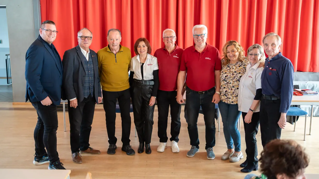 Sieben Personen stehen zusammen auf einem Holzboden vor einem roten Vorhang und lächeln für ein Foto. Die beiden Männer an den Enden tragen eine Brille und Lederschuhe. Die Frau in der Mitte trägt ein weißes Hemd und schwarze Hose. Die anderen drei Männer tragen rote Polo-Shirts.