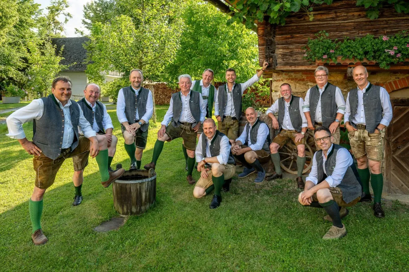 Eine Gruppe von Männern in traditioneller Kleidung posiert für ein Foto vor einem rustikalen Haus, einige sitzen auf einem Fass.