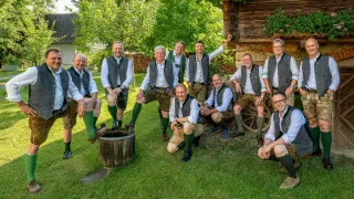 Eine Gruppe von Männern in traditioneller Kleidung posiert für ein Foto vor einem rustikalen Haus, einige sitzen auf einem Fass.
