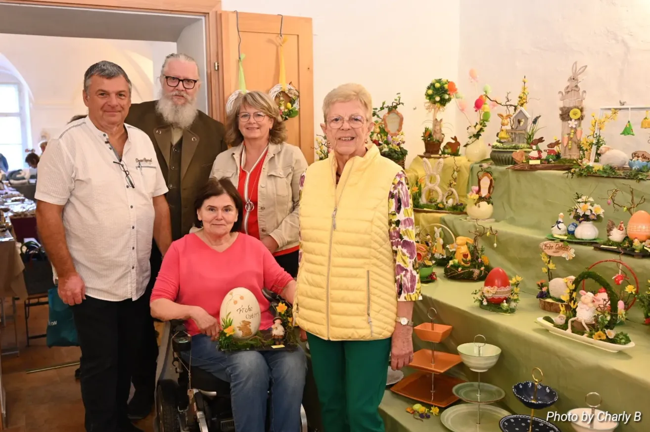 Eine Gruppe von Menschen steht um einen Tisch mit Osterdekoration. Eine ältere Frau im Rollstuhl hält ein bemaltes Ei. Eine Frau in einer gelben Weste steht neben ihr.