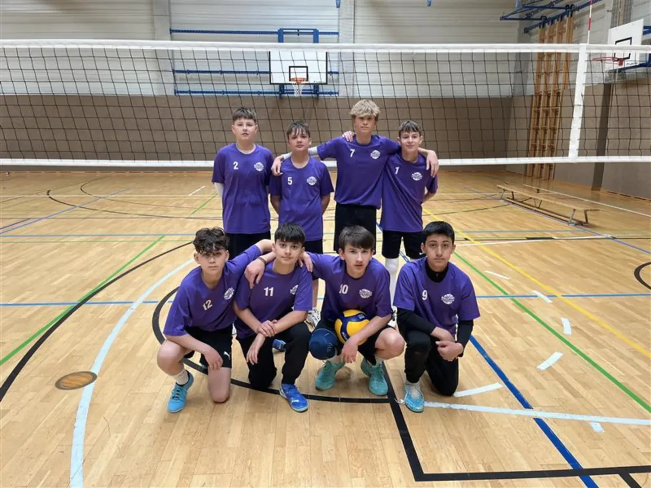 Sieben junge Männer in lila Trikots posieren für ein Foto in einer Turnhalle mit einem Volleyballnetz im Hintergrund.
