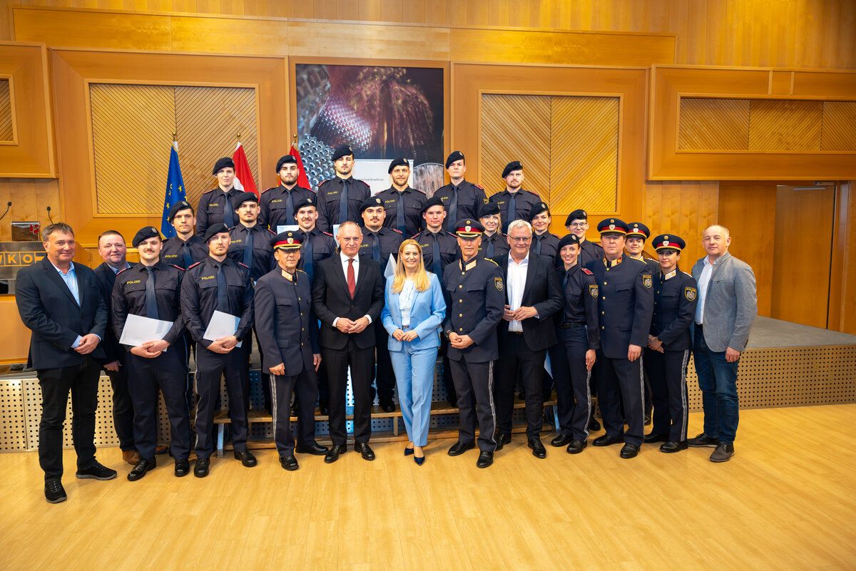 Eine Gruppe uniformierter Personen steht in Formation, flankiert von zwei Männern und einer Frau vorne. Es handelt sich wahrscheinlich um Polizeibeamte, die für ein Gruppenfoto posieren.