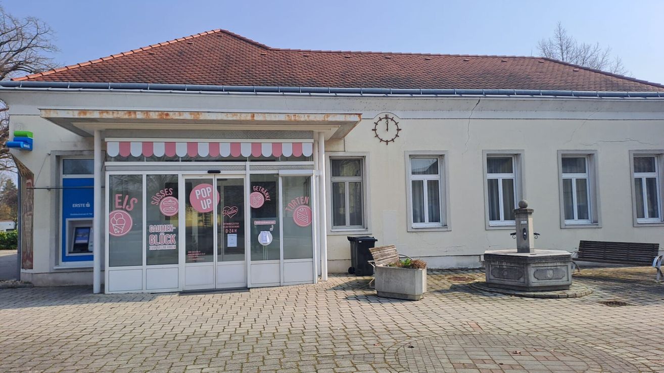 Ein weißes Gebäude mit rotem Dach hat Glastüren mit den Texten POP UP und SÜSSES GAUME GLÜCK. Ein Mülleimer steht vor der Tür. Eine Bank mit einem Topf steht rechts. Eine Uhr ist an der Wand über den Fenstern montiert.