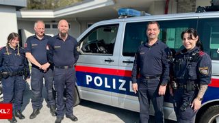 Vier Polizeibeamte stehen vor einem silbernen Polizeiwagen mit 'POLIZEI' an der Seite. Der Wagen hat blaue, rote und weiße Streifen.