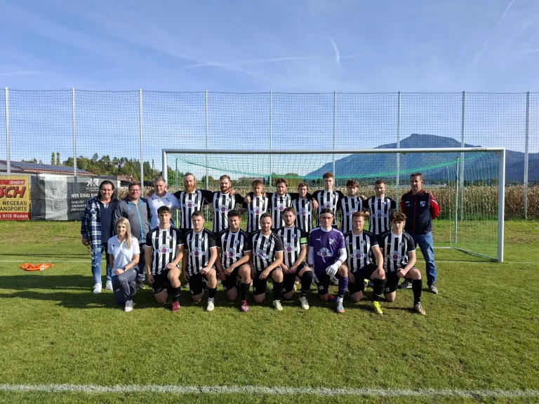 Eine Fußballmannschaft posiert für ein Foto vor einem Tornetz mit Bergen im Hintergrund. Sie tragen schwarze und weiße Trikots.
