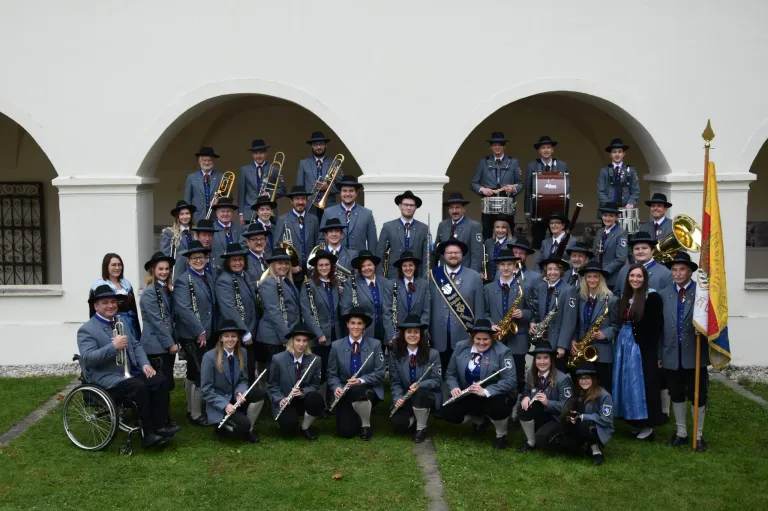 Eine Gruppe von Musikern in einheitlichen Kostümen posiert für ein Gruppenfoto vor einem Bogen. Sie halten Instrumente wie Trompeten, Posaunen, Trommeln und Flöten.