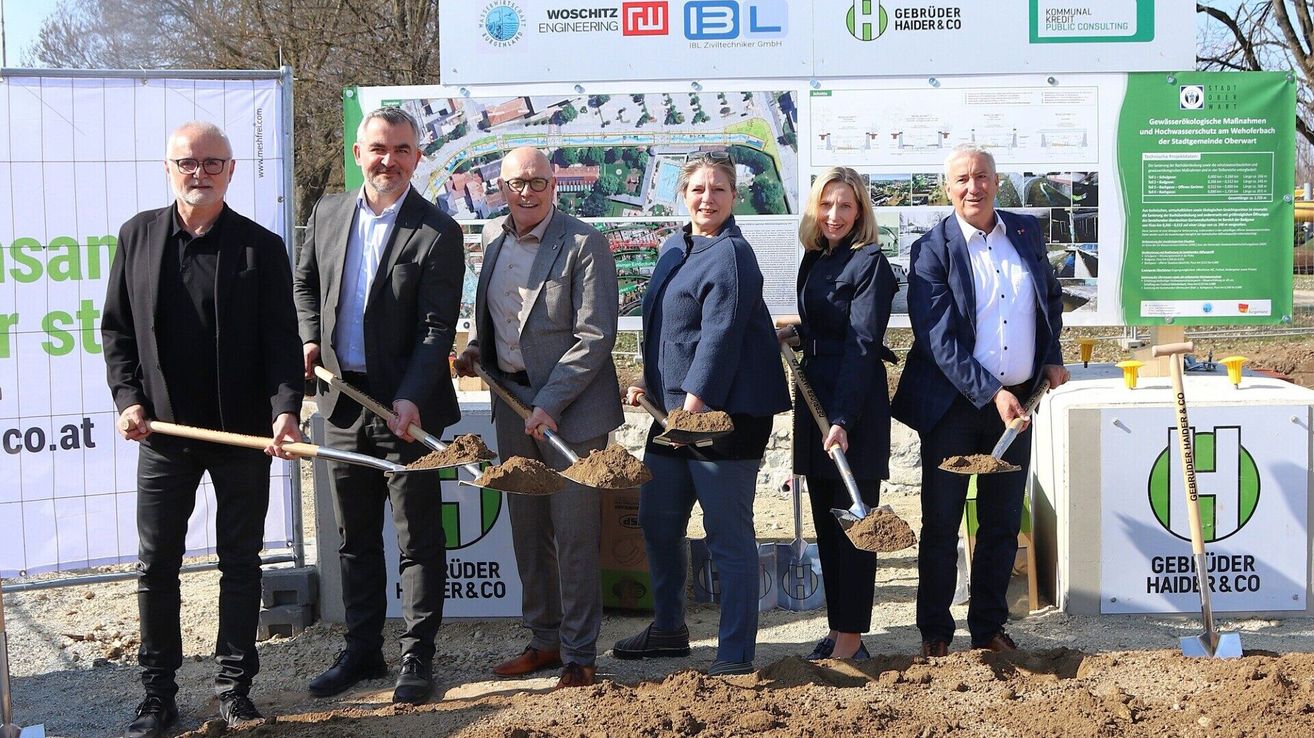 Fünf Personen in formeller Kleidung halten Schaufeln vor einer Baustelle und lächeln für die Kamera. Dahinter zeigt ein Banner verschiedene Logos und eine Karte.