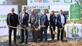 Fünf Personen in formeller Kleidung halten Schaufeln vor einer Baustelle und lächeln für die Kamera. Dahinter zeigt ein Banner verschiedene Logos und eine Karte.