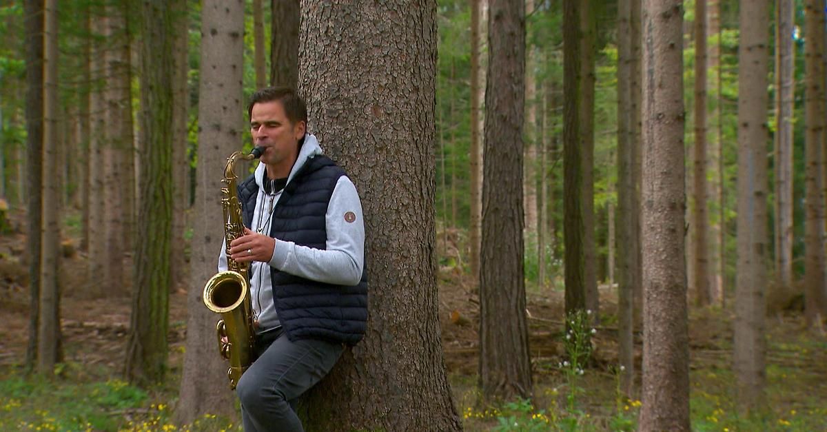 Ein Mann steht in einem Wald, lehnt sich gegen einen Baum und spielt Saxophon. Er trägt eine weiße Jacke und eine blaue Weste.