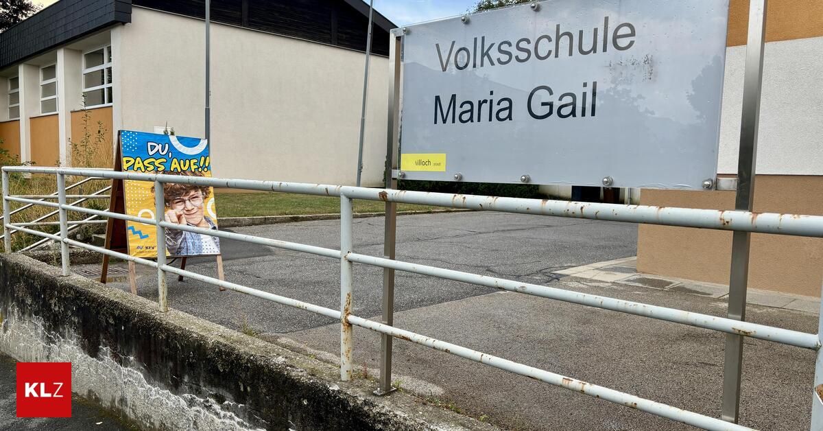 Ein weißes Schild mit der Aufschrift 'Volksschule Maria Gail' ist an einem Metallzaun angebracht. Daneben befindet sich ein Poster mit dem Gesicht einer Frau und dem Text 'AUE!'. Im Hintergrund steht ein Gebäude mit einer Ziegelsteinmauer.