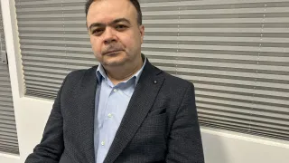 Ein Mann in einem dunklen Anzug mit blauem Hemd und schwarzem Einstecktuch sitzt in einem Büro.