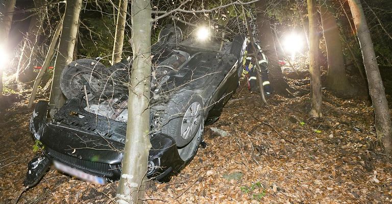Ein beschädigtes schwarzes Auto liegt auf der Seite im Wald, mit einem Feuerwehrmann in der Nähe. Die Front des Autos ist zerquetscht und die Lichter sind an.