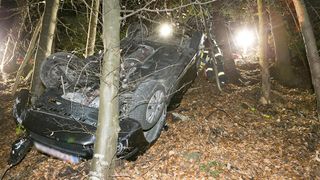 Ein beschädigtes schwarzes Auto liegt auf der Seite im Wald, mit einem Feuerwehrmann in der Nähe. Die Front des Autos ist zerquetscht und die Lichter sind an.
