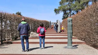 Eine Gruppe von Menschen geht eine Treppe hinauf. Ein Mann und eine Frau stehen im Vordergrund, während andere weiter oben sind. Die Umgebung ist im Freien mit Bäumen und Büschen im Hintergrund.