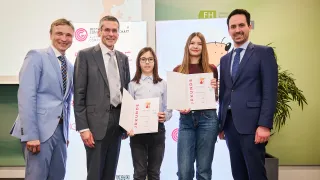 Fünf Erwachsene und zwei junge Mädchen, eines mit Brille und das andere mit langen Haaren, stehen zusammen und halten Urkunden. Sie sind alle in formeller Kleidung. Hinter ihnen ist ein Banner mit einem Cartoon-Charakter zu sehen.