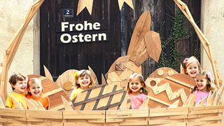 Vier lächelnde junge Mädchen stehen vor einer hölzernen Dekoration mit Ostereiern und einem Schild, das Frohe Ostern steht.