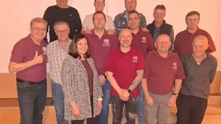 Eine Gruppe von Menschen, einschließlich Männern und Frauen, posiert für ein Foto. Sie tragen alle rote Hemden mit einem Logo. Einige von ihnen tragen Brillen. Sie alle lächeln und scheinen für ein Gruppenfoto zu posieren.