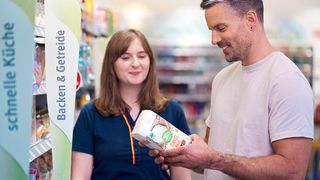 Ein Mann und eine Frau sind in einem Geschäft. Der Mann lächelt und zeigt der Frau ein Produkt. Die Frau schaut ihn an. Hinter ihnen steht ein Schild, das 'Backen & Getreide' sagt.