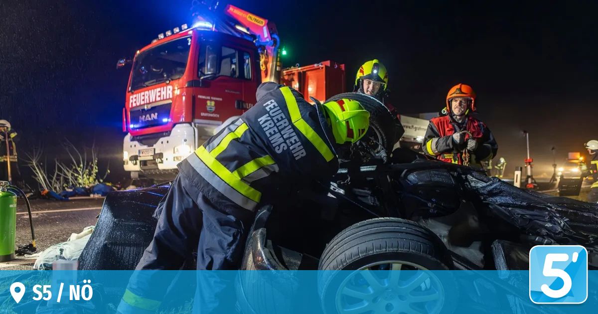 Feuerwehrleute in Schutzkleidung arbeiten nachts an einem demolierten Fahrzeug. Dahinter steht ein roter Feuerwehrwagen mit blinkenden Lichtern.