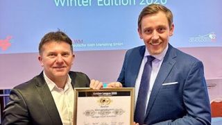 Zwei formell gekleidete Männer posieren für ein Foto mit einer Plakette. Die Plakette lautet 'Goldene Liga 2020'. Sie lächeln und scheinen sich bei einer Veranstaltung zu befinden.