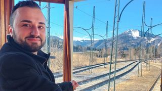Ein Mann mit Bart schaut aus dem Zugfenster und zeigt eine Aussicht auf eine Eisenbahnstrecke mit Bergen im Hintergrund.
