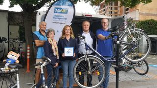 Fünf Personen stehen mit Fahrrädern, eine hält eine Schachtel, unter einem Banner mit der Aufschrift MoMa und Mobilitäts Management. Sie stehen auf einem Bürgersteig in der Nähe eines Lieferwagens.