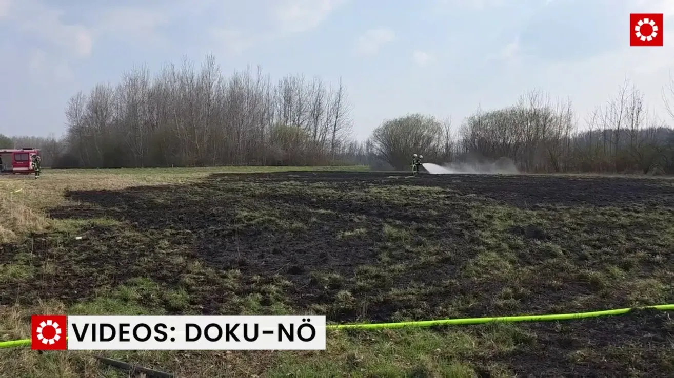 Zwei Feuerwehrleute löschen ein Feuer in einem Feld. Das Feld ist größtenteils kahl mit einigen Grasbüscheln. Bäume sind im Hintergrund sichtbar.