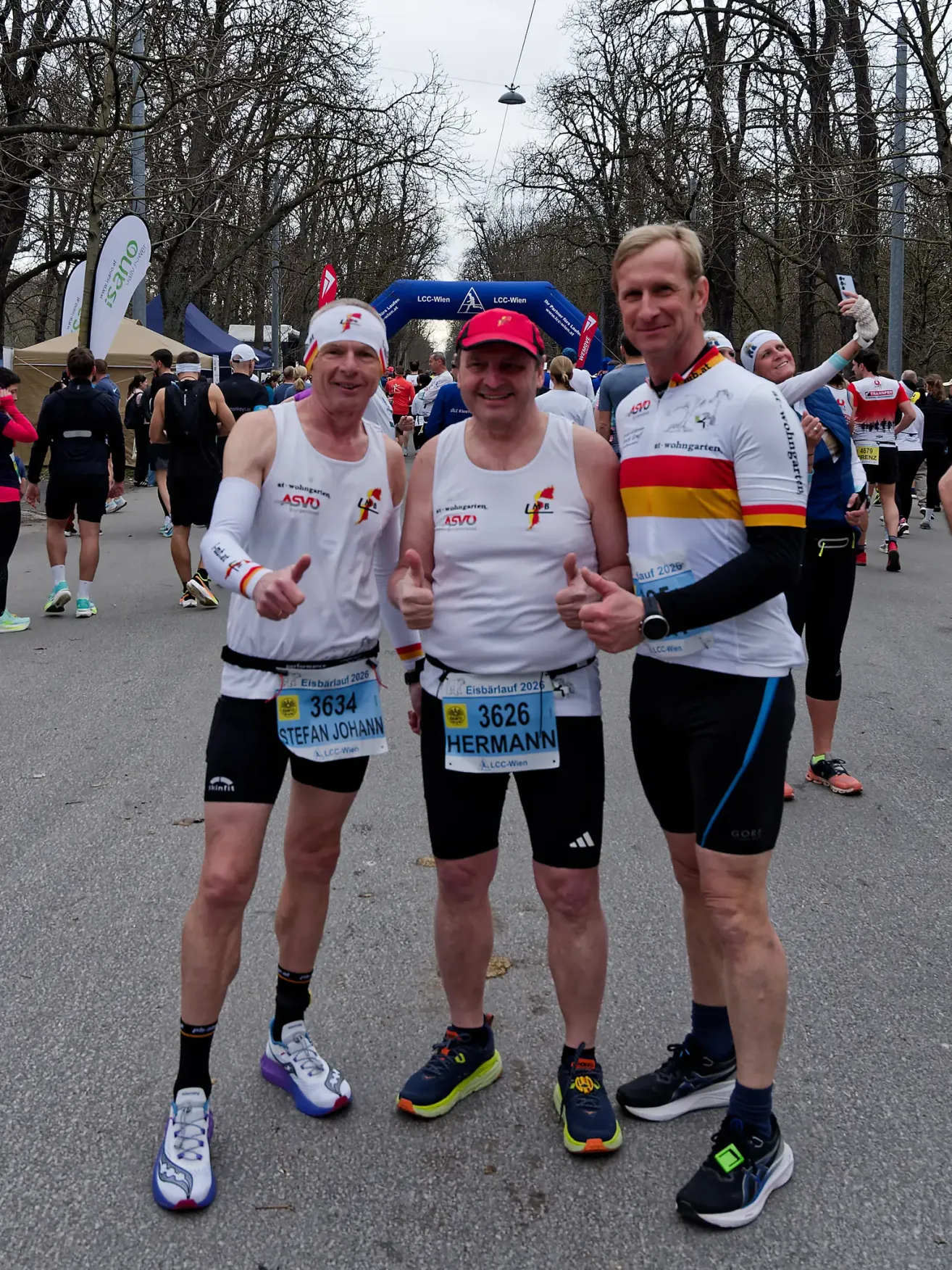 Drei Männer posieren für ein Foto während eines Marathons, tragen weiße Shirts, Mützen und Nummernschilder. Hinter ihnen gehen eine Menge anderer Läufer und eine Frau hält ein Telefon. Bäume und Banner sind im Hintergrund sichtbar.