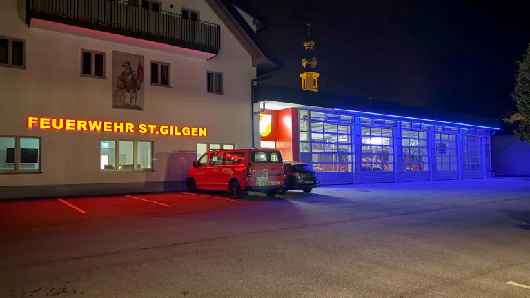 Ein roter Van und ein schwarzes Auto sind vor einem Gebäude geparkt. Das Gebäude hat die Aufschrift HR ST. GILNGEN auf der Vorderseite.
