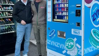 Zwei Männer stehen vor einem blauen Verkaufsautomaten, einer trägt eine Brille und Turnschuhe, der andere eine Jacke und Turnschuhe. Der Automat bietet Produkte wie ZYN und Marlboro an.