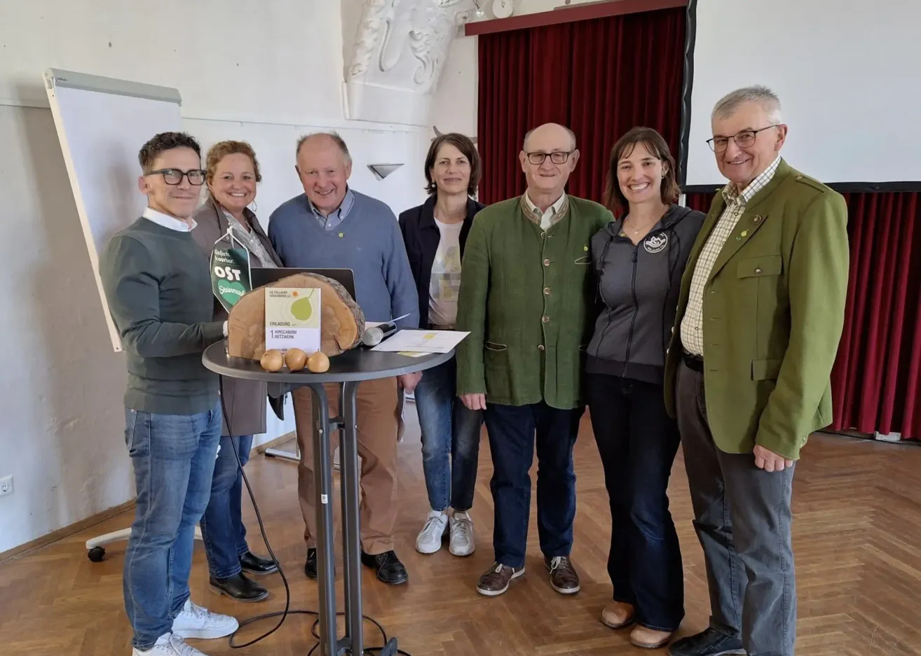 Eine Gruppe von Menschen steht um einen Tisch mit einem apfelförmigen Objekt, möglicherweise zur Feier eines Ereignisses. Der Mann links trägt eine Brille und einen grauen Pullover.
