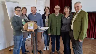 Eine Gruppe von Menschen steht um einen Tisch mit einem apfelförmigen Objekt, möglicherweise zur Feier eines Ereignisses. Der Mann links trägt eine Brille und einen grauen Pullover.