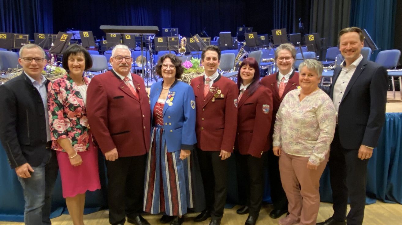 Sechs Personen in formeller Kleidung stehen auf einem Holzboden. Dahinter befinden sich Musikinstrumente, was auf einen Aufführungsort hindeutet.